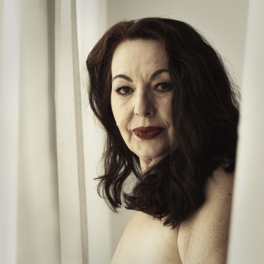 Frau mit langen, dunklen Haaren, schaut ernst durch einen Vorhang.