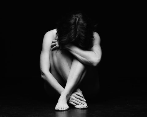 Eine sitzende Person mit angezogenen Knien, den Kopf in den Händen, in dunkler Umgebung.