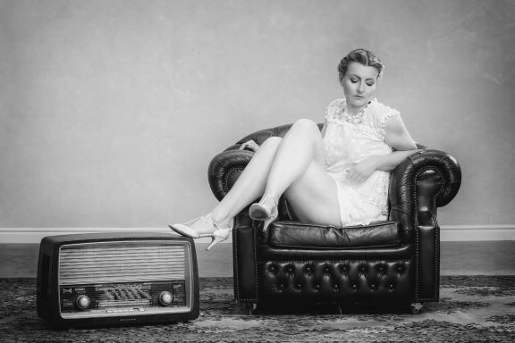 Eine Frau in einem weißen Kleid sitzt auf einem Chesterfield-Sofa neben einem alten Radio.