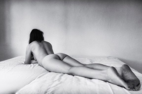 Akt einer Person, die auf einem Bett liegt, Rückenansicht. Schwarz-weiß Fotografie.